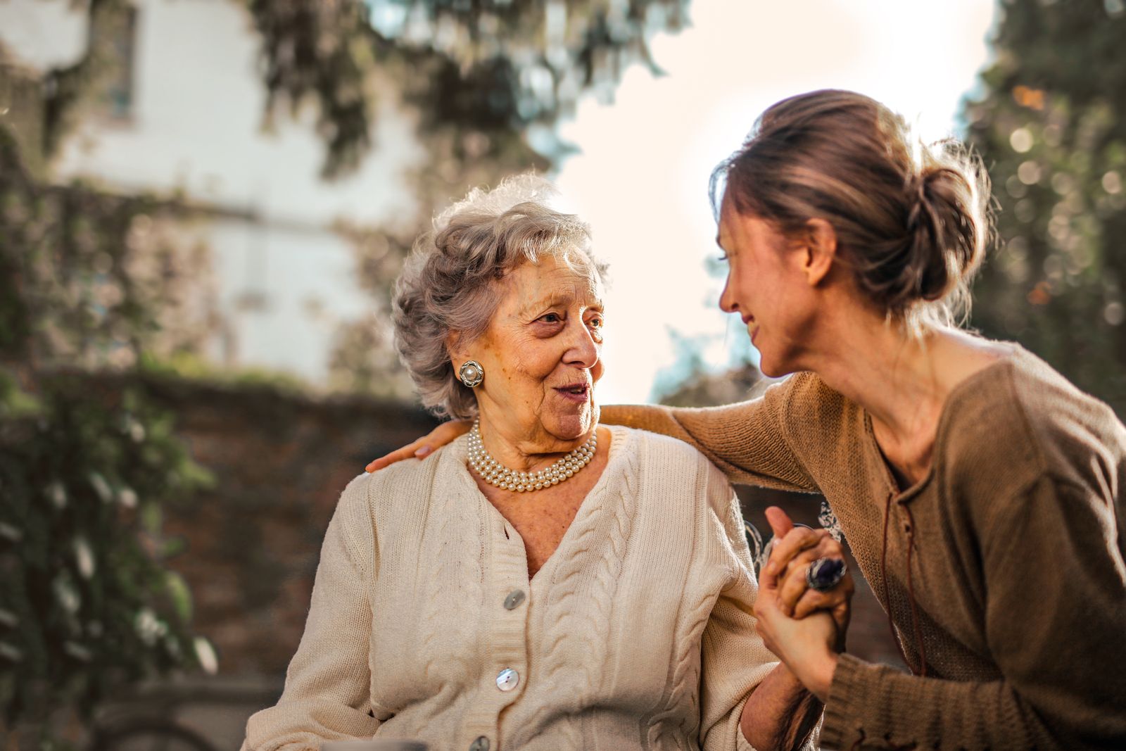 An elderly woman in a pearl necklace sharing a warm conversation with a younger woman in a sunny garden