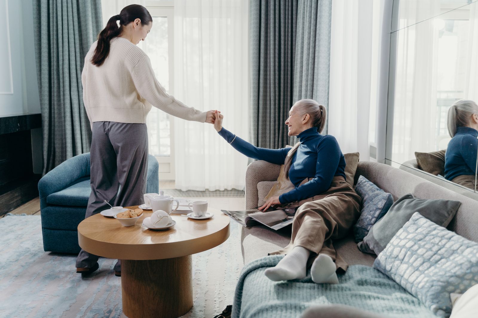 An adult daughter warmly helping her elderly mother up from the sofa in a bright living room