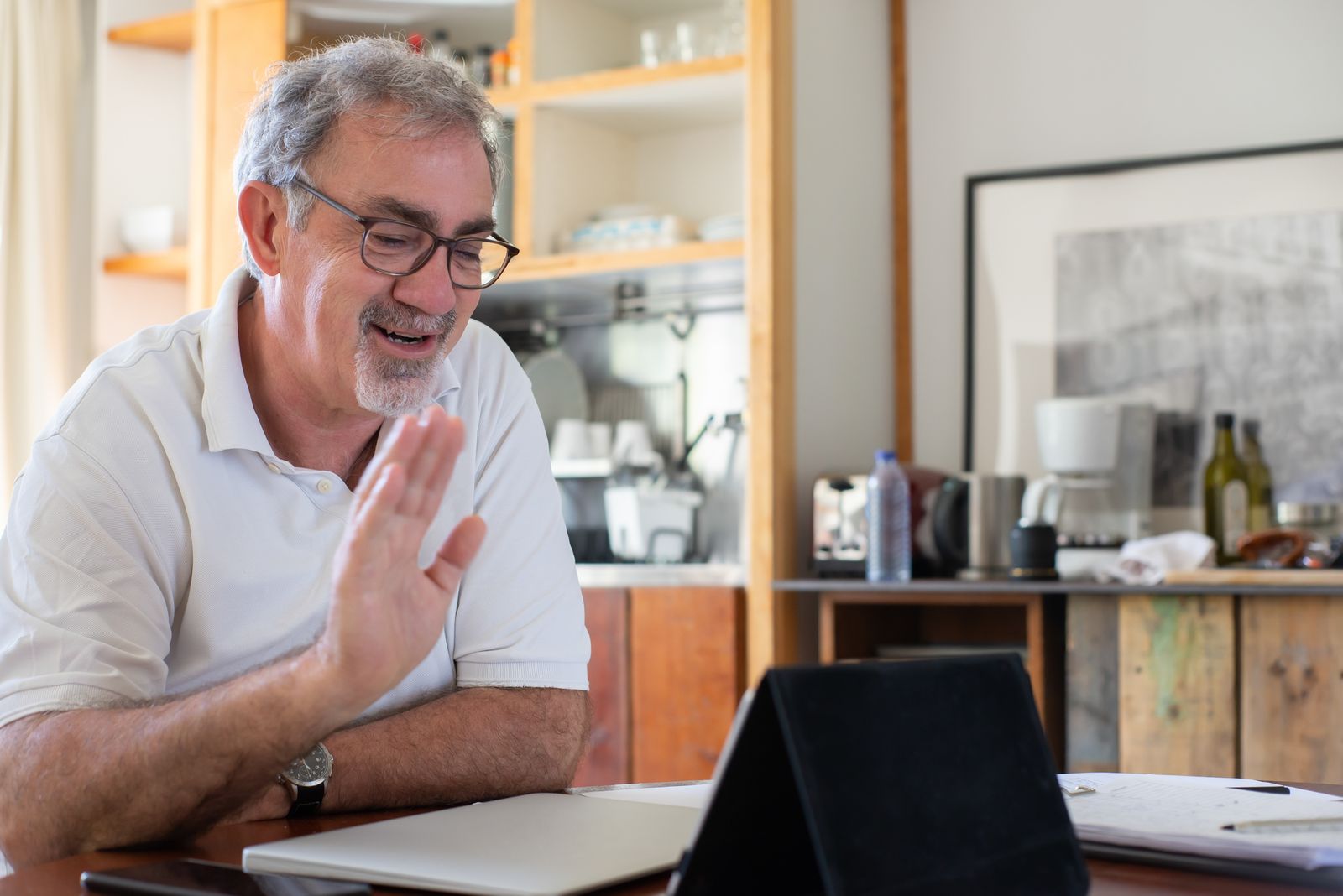 A smiling older man waving during a video call on his tablet in a cozy home kitchen